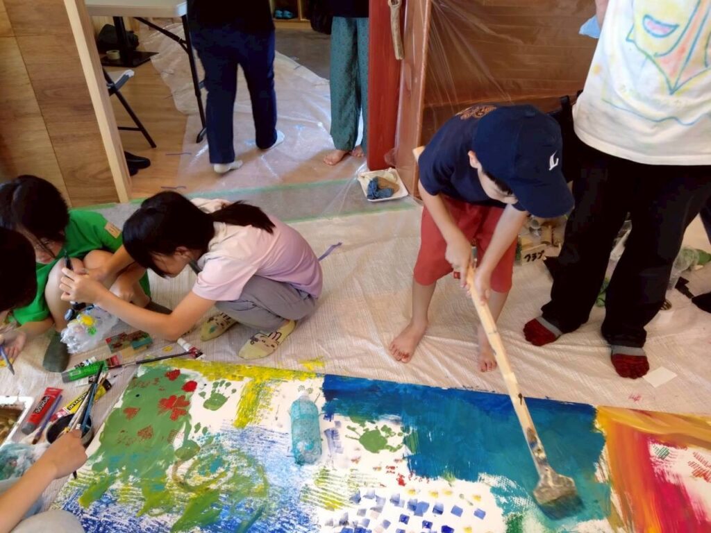 大きな板に子供たちが刷毛や筆を使って元気に絵を描いている様子