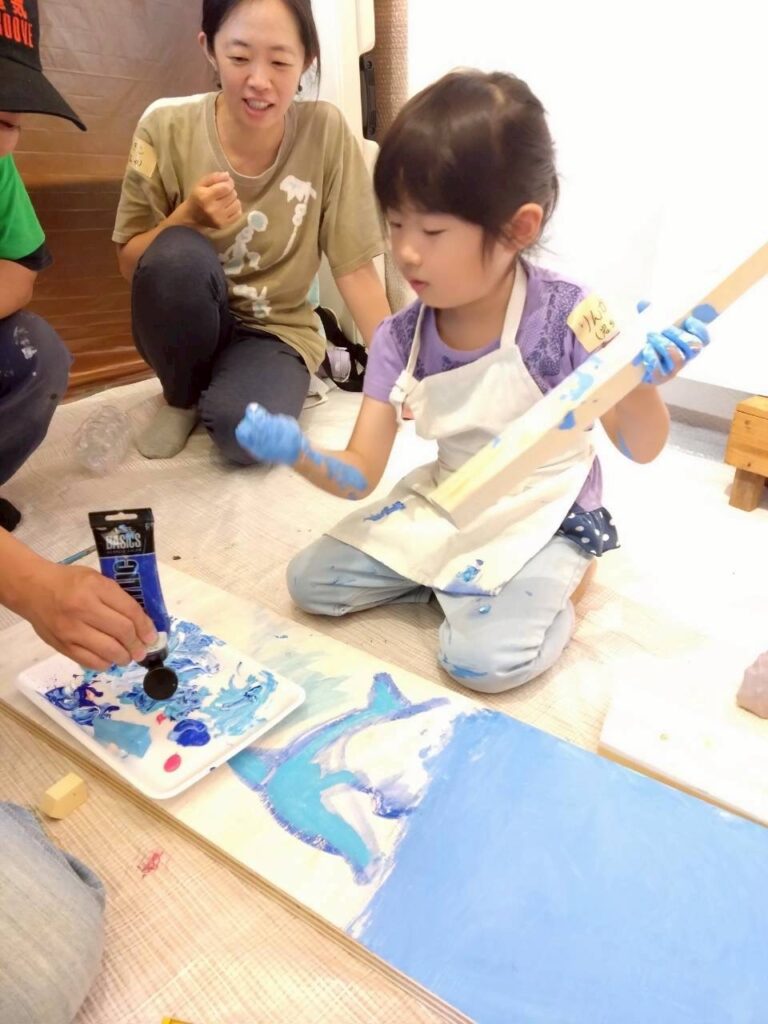 木のパーツを持って大人に絵の具を追加してもらう子供の様子
