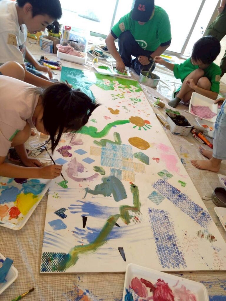 大きな板に子供たちが絵の具で各々模様を描いている風景